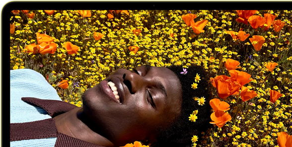 MacBook Neo, citrus colour, screen shows a smiling person lying in a sunny field of flowers
